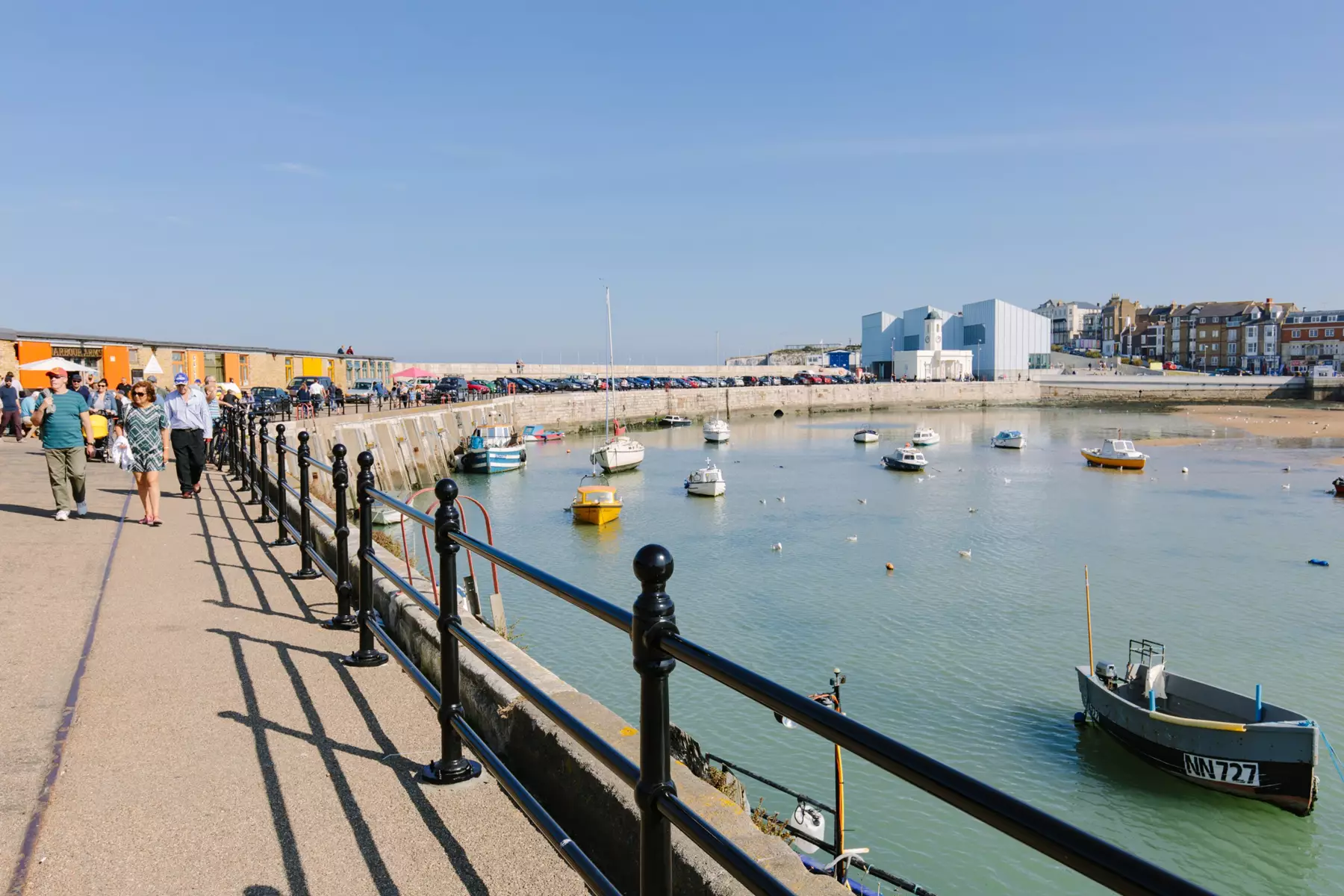 People walking along stone pier. Bright blue sky. Tide in with boats in the harbour. Gallery and residential buildings at the start of the pier